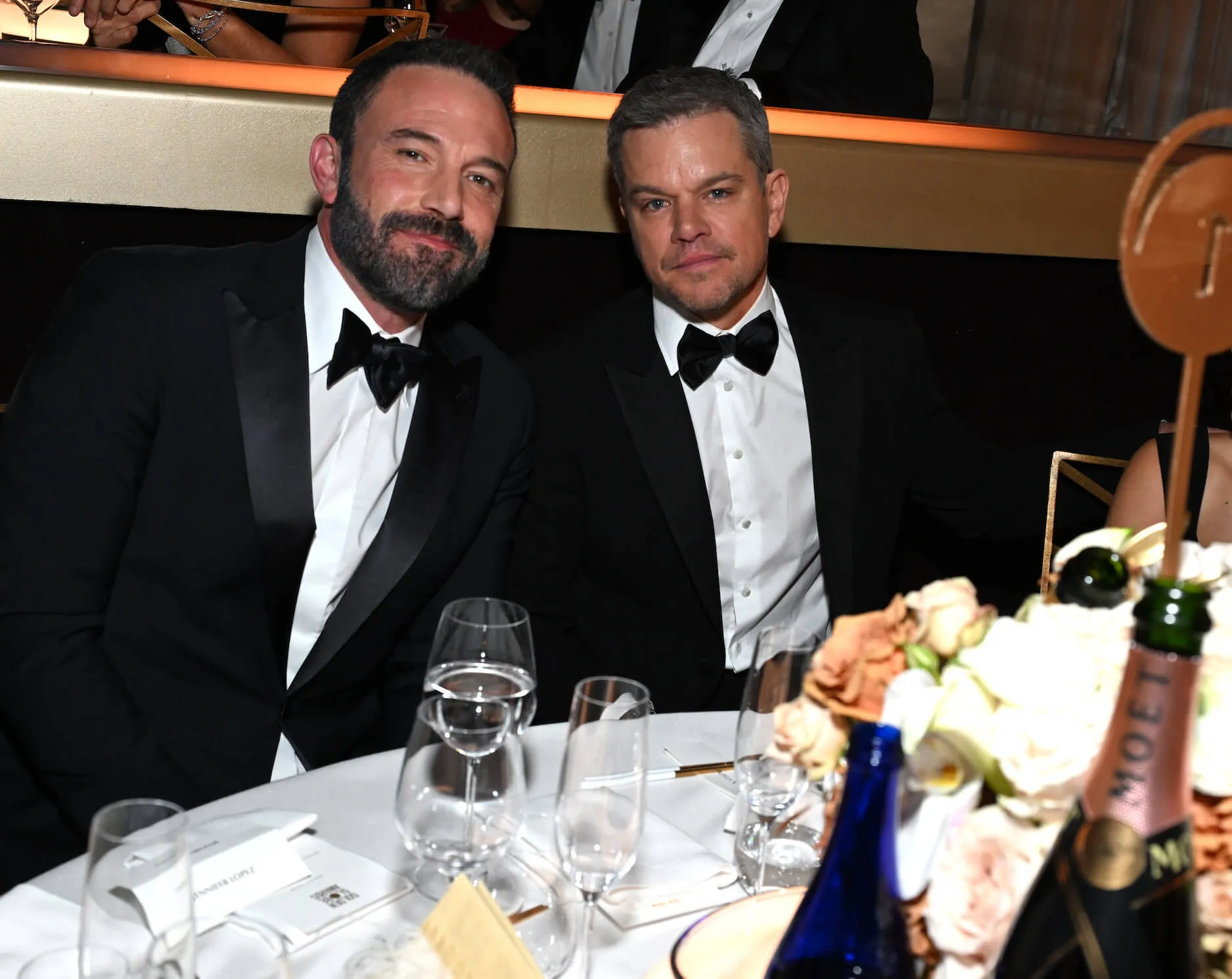 Ben Affleck and Matt Damon smiling while sitting next to each other at the 81st Annual Golden Globes. They are both wearing tuxedos and black bow ties.