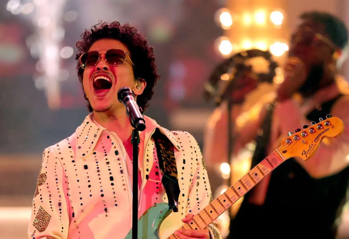 Bruno Mars wears sunglasses and holds a guitar while he sings into a microphone.