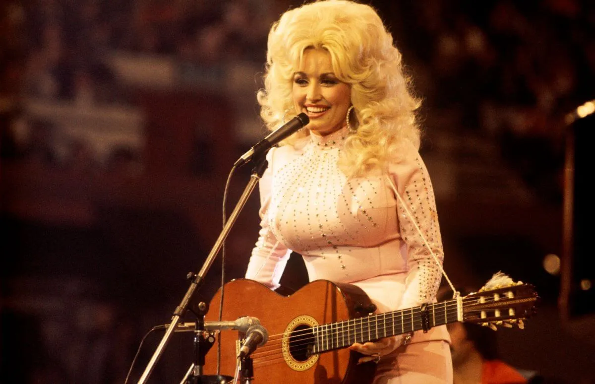 Dolly Parton wears a white turtleneck and stands in front of a microphone with a guitar.