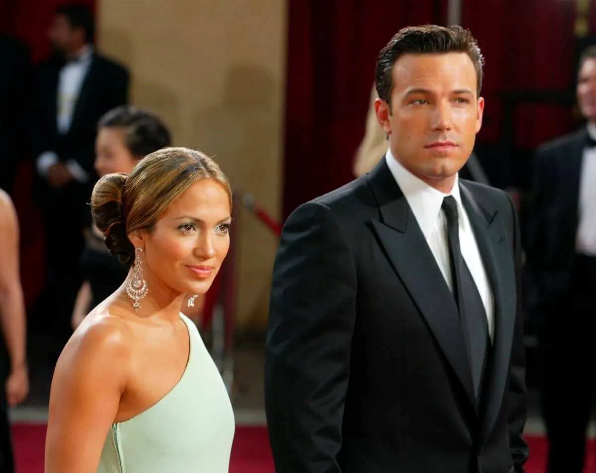 Jennifer Lopez wears a teal one-shoulder dress and stands next to Ben Affleck, who wears a suit.