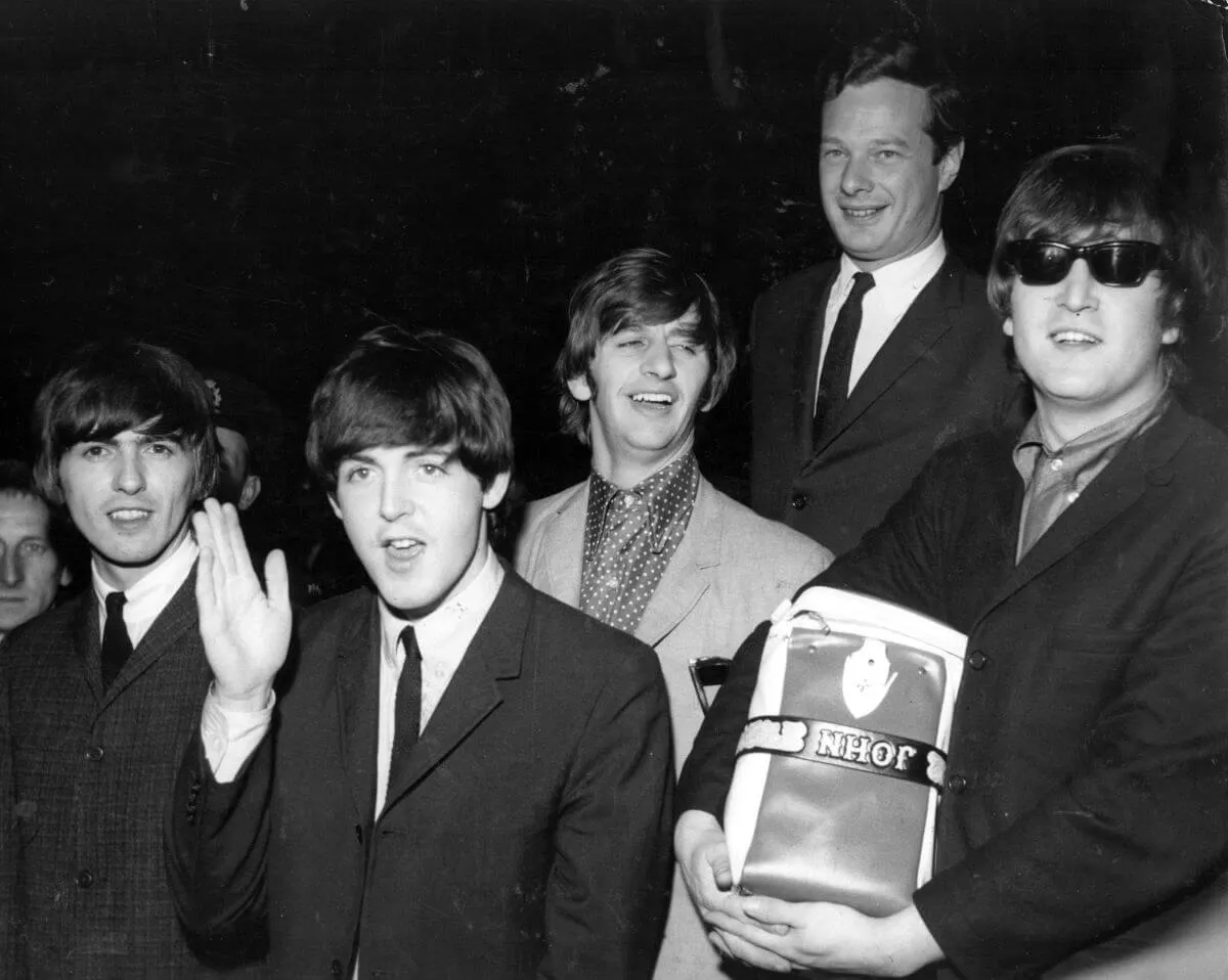 A black and white picture of The Beatles and Brian Epstein wearing suits at the airport. John Lennon wears sunglasses and holds a suitcase.