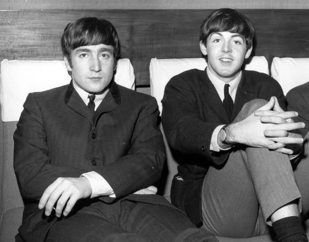 A black and white picture of John Lennon and Paul McCartney sitting on a couch. They both wear suits.