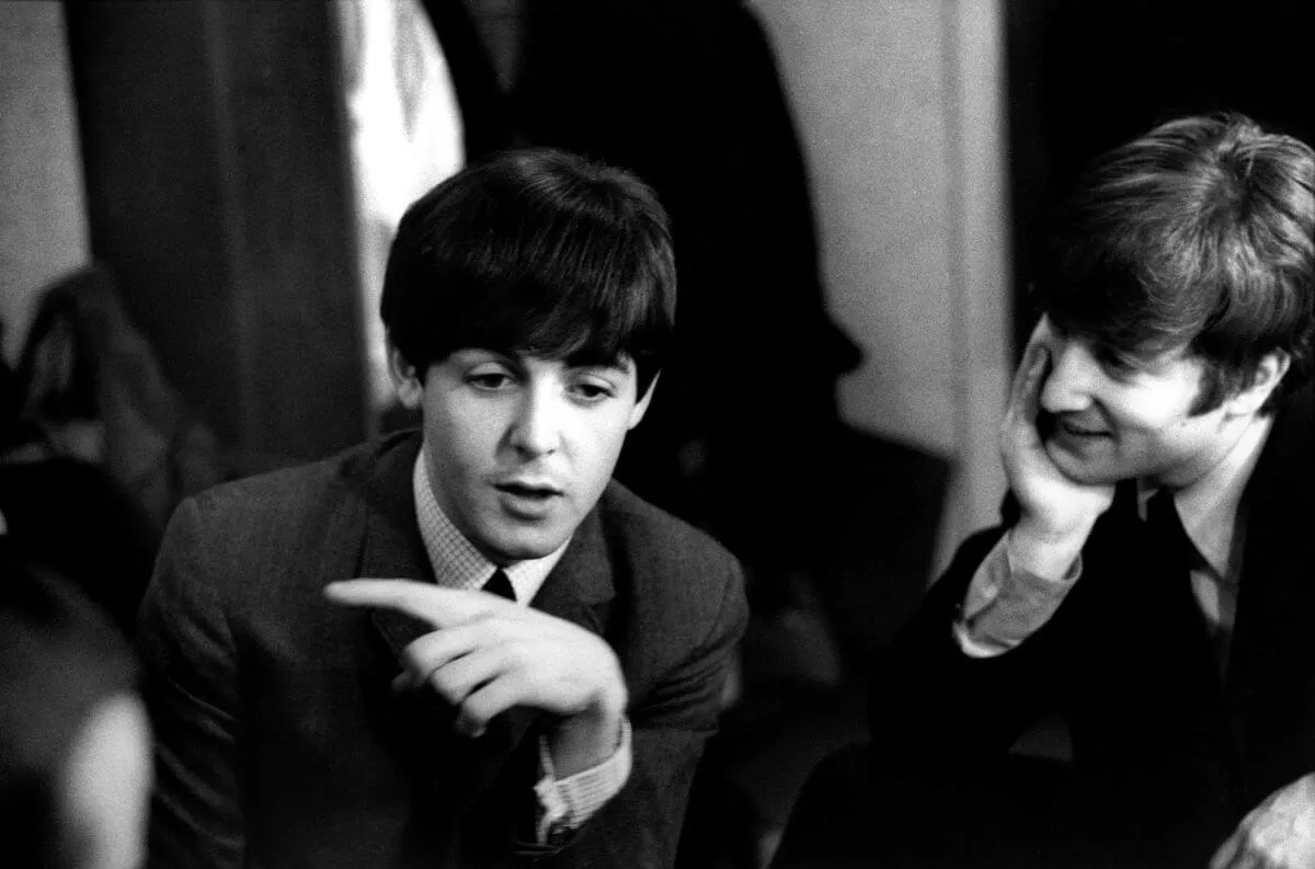 A black and white picture of Paul McCartney and John Lennon wearing suits. Lennon watches McCartney as he speaks.