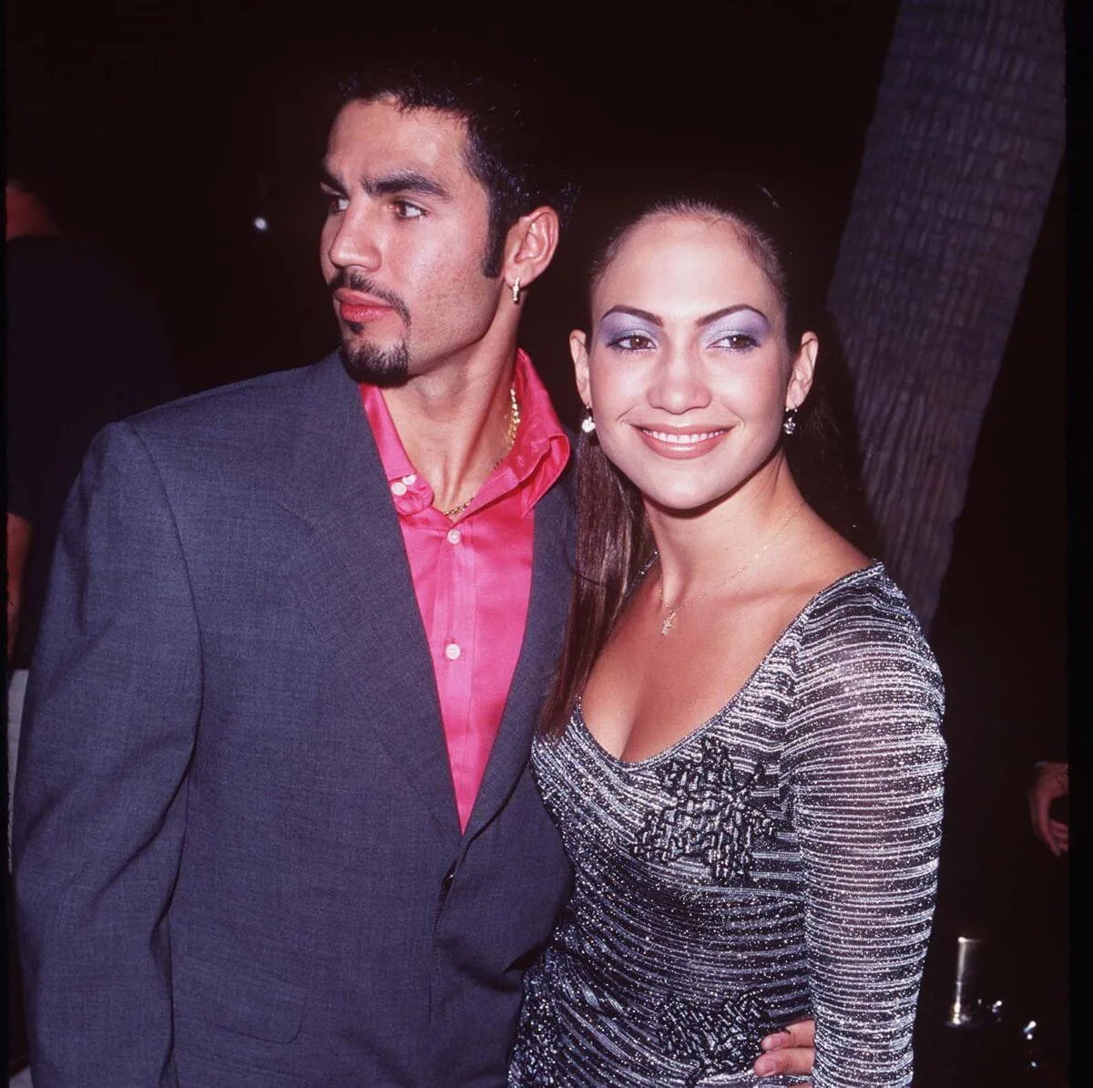 Ojani Noa wears a pink shirt and gray jacket and stands with Jennifer Lopez, who wears a silver dress.