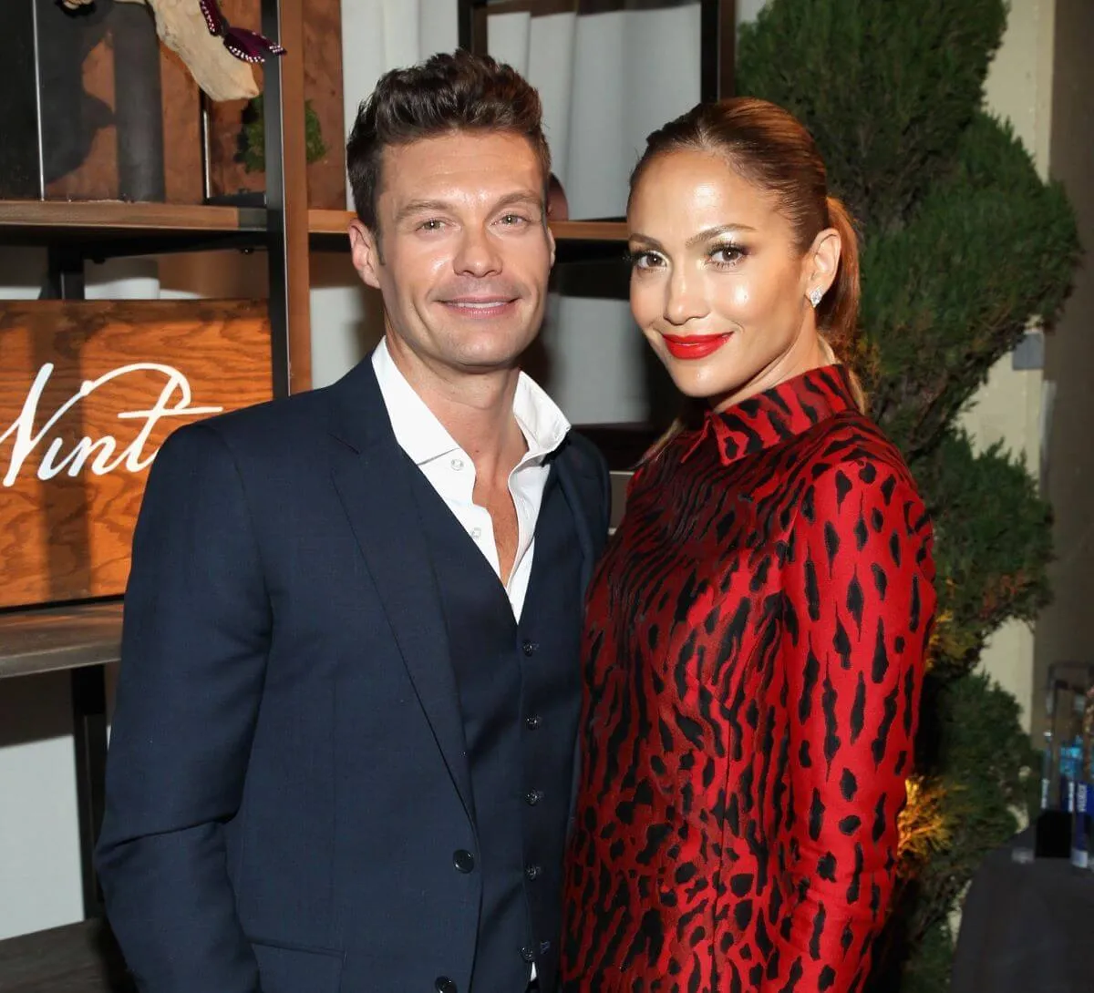 Ryan Seacrest wears a blue suit and stands next to Jennifer Lopez, who wears a red dress with black spots.